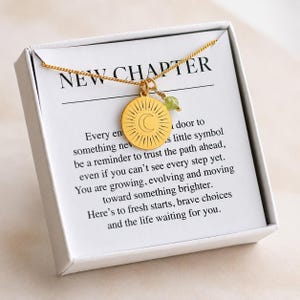 Pode incluir: Um colar dourado com um pingente circular com uma gravação de sol e lua crescente, acompanhado por uma pequena conta verde. O colar é exibido num cartão branco com o texto NEW CHAPTER e uma mensagem inspiradora.