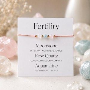 Puede incluir: Una delicada pulsera con cuentas de piedra lunar, cuarzo rosa y aguamarina. La pulsera está ensartada en un fino cordón rosa claro y adornada con cuentas doradas. La tarjeta detrás de la pulsera dice "Fertilidad".