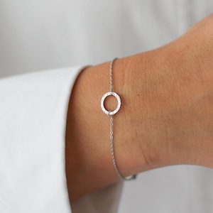 May include: A silver bracelet with a textured circular pendant. The bracelet is attached to a fine chain. The design is minimalist and elegant. The silver colour gives it a classic and versatile appeal. The bracelet is shown on a wrist.