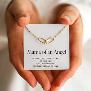 Puede incluir: Un collar dorado con dos colgantes ovalados entrelazados se muestra en una tarjeta blanca. La tarjeta dice "Mama of an Angel" con un mensaje sentimental debajo. El collar se sostiene en manos abiertas.