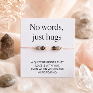 May include: A bracelet featuring grey, brown, and white beads on a pink cord, displayed on a white card. The card reads "No words, just hugs" and a message about love. Crystals and flowers are in the background, creating a soft aesthetic.