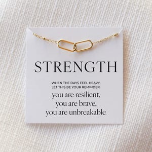 May include: A gold-coloured necklace featuring two interlocking oval links, presented on a white card. The card displays the word "STRENGTH" and an inspirational message: "you are resilient, you are brave, you are unbreakable."