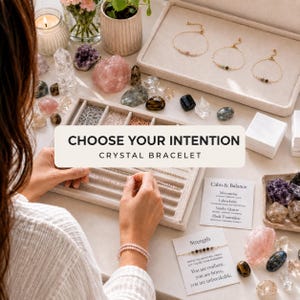 Puede incluir: Una colección de delicadas pulseras de cristal exhibidas en una bandeja de terciopelo beige, acompañadas de piedras preciosas en bruto, una vela y tarjetas informativas con el texto CHOOSE YOUR INTENTION CRYSTAL BRACELET, Calm & Balance y Strength.