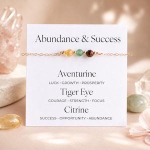 May include: A gold chain bracelet featuring three gemstone beads: yellow citrine, green aventurine, and brown tiger eye. The bracelet is presented in front of a white card with the words "Abundance & Success" and gemstone descriptions.
