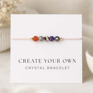 May include: A crystal bracelet with orange, gray, black, purple, and gray beads strung on a pink cord. The bracelet is displayed on a white card with the text "CREATE YOUR OWN CRYSTAL BRACELET" in black.