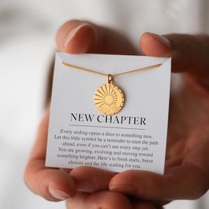 Könnte beinhalten: Eine goldfarbene Halskette mit einem runden Anhänger, der Sonne und Mond zeigt. Der Anhänger hängt an einer feinen Kette, präsentiert auf einer weißen Karte mit dem Text "NEW CHAPTER" und inspirierenden Worten.