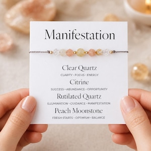 May include: A gemstone bracelet featuring clear quartz, citrine, rutilated quartz, and peach moonstone. The bracelet is presented on a card with the word "Manifestation" and stone properties. The bracelet is held by two hands.