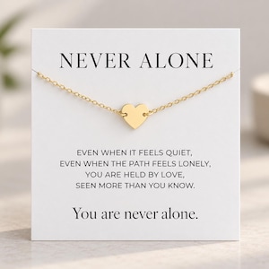 May include: A gold-tone heart pendant necklace displayed on a white card featuring the text NEVER ALONE at the top, followed by the message: EVEN WHEN IT FEELS QUIET, EVEN WHEN THE PATH FEELS LONELY, YOU ARE HELD BY LOVE, SEEN MORE THAN YOU KNOW.