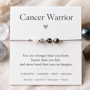 May include: A Cancer Warrior bracelet with a gray cord and a series of gemstone beads. The card behind the bracelet has the words "Cancer Warrior" and an inspirational message about strength and courage.