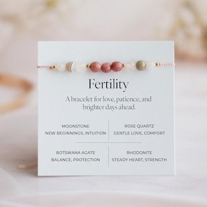 May include: A delicate bracelet with pink and neutral-toned beads, strung on a thin, light pink cord. The bracelet is displayed on a white card with the word "Fertility" and text about love, patience, and brighter days. The card lists the stones used.