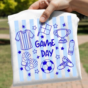 May include: A white t-shirt with light blue vertical stripes featuring a blue graphic print that says GAME DAY. The design includes illustrations of a soccer jersey, a referee flag, a trophy, a water bottle, goalkeeper gloves, a soccer ball, and cleats.