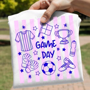 May include: A white t-shirt with vertical light pink stripes and a blue graphic print featuring the text "GAME DAY" surrounded by illustrations of a soccer jersey, a referee flag, a trophy, a water bottle, soccer cleats, gloves, and a ball.