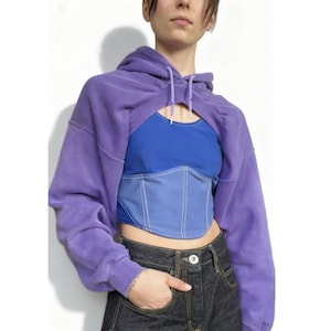 Könnte beinhalten: Ein lila Crop-Hoodie mit Kapuze und Kordelzug, getragen über einem blauen Oberteil und einem hellblauen Korsett mit weißen Nähten. Das Model trägt eine dunkelblaue Jeans.