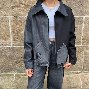May include: A two-tone denim jacket with a black and gray color scheme. The jacket features a zipper closure, a collar, and two front pockets. The left pocket has a black embroidered "R" design. The jacket is paired with matching denim jeans.