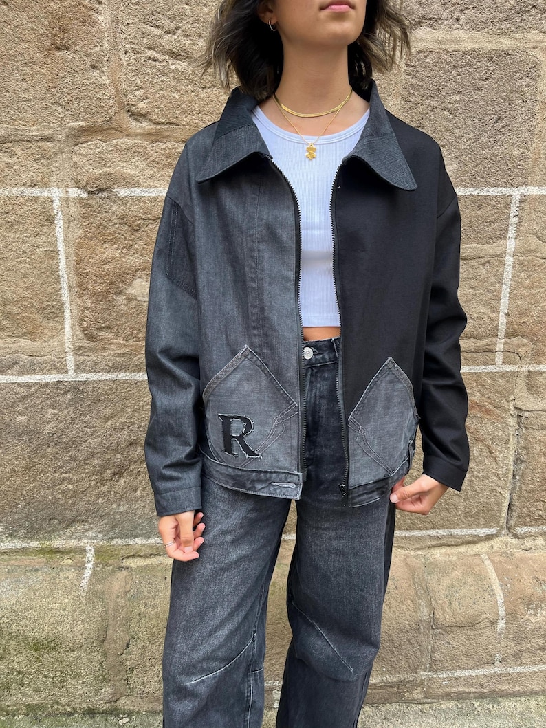 May include: A two-tone denim jacket with a black and gray color scheme. The jacket features a zipper closure, a collar, and two front pockets. The left pocket has a black embroidered "R" design. The jacket is paired with matching denim jeans.