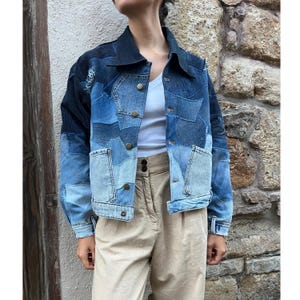 Op de afbeelding: Een patchwork spijkerjasje in verschillende blauwtinten. De jas heeft een kraag, knopen en voorzakken. De mouwen zijn opgerold en de jas wordt gedragen over een witte top en een beige broek.