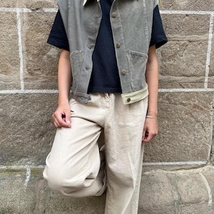 May include: A gray vest with a cream-colored collar and trim, worn over a black t-shirt. The vest has button closures and pockets. The person is wearing beige pants. The background is a textured stone wall.