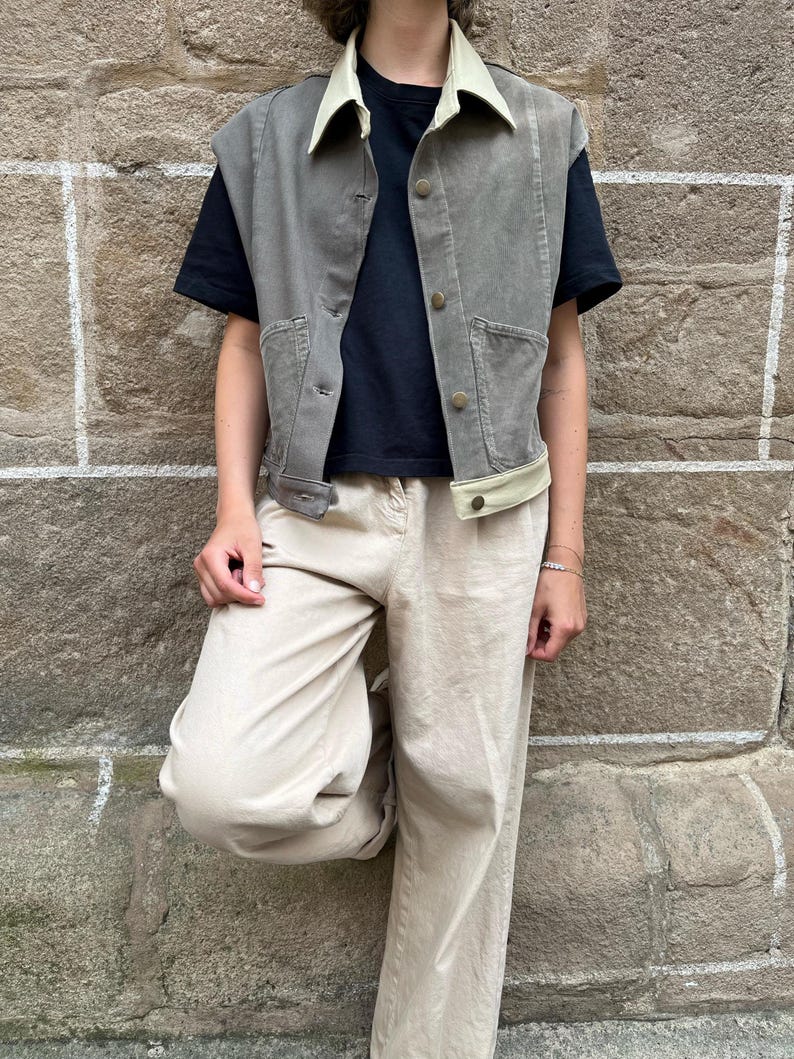 May include: A gray vest with a cream-colored collar and trim, worn over a black t-shirt. The vest has button closures and pockets. The person is wearing beige pants. The background is a textured stone wall.
