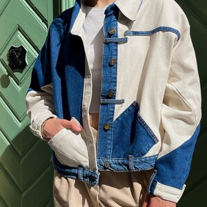 May include: A two-tone denim jacket with a white and blue color block design. The jacket features a classic collar, button closures, and front pockets. The sleeves are long, and the jacket is worn open. The jacket is a stylish outerwear piece.