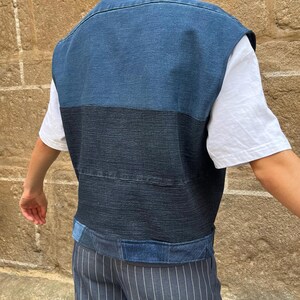 May include: A denim vest in shades of blue, with a contrasting dark blue horizontal stripe across the midsection. The vest has a collared neckline and appears to be worn over a white t-shirt. The garment is part of a clothing collection.