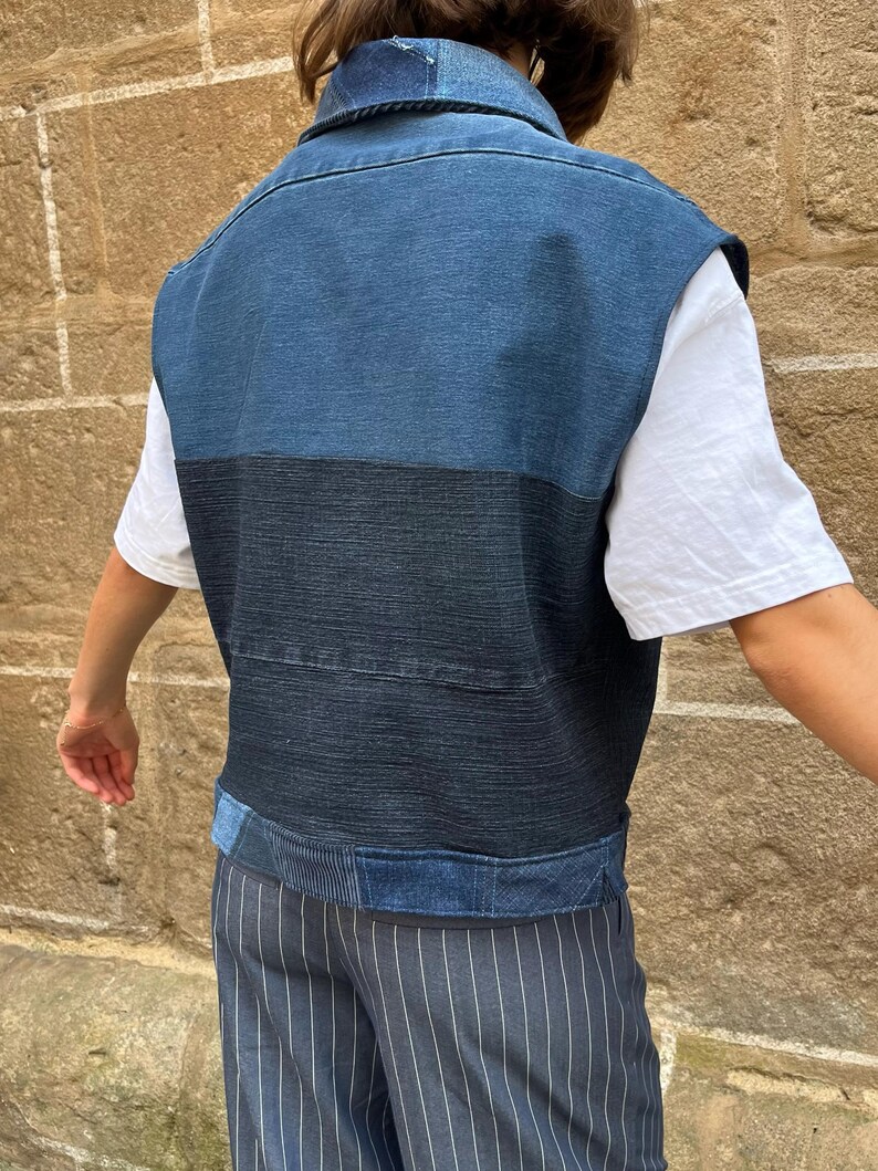 May include: A denim vest in shades of blue, with a contrasting dark blue horizontal stripe across the midsection. The vest has a collared neckline and appears to be worn over a white t-shirt. The garment is part of a clothing collection.