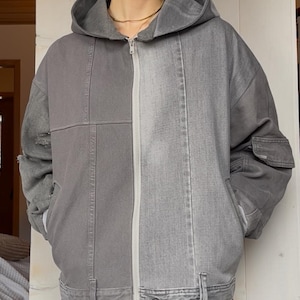 May include: A gray hooded jacket with a zip-up front. The jacket is a patchwork design, with different shades of gray denim. It has a hood, pockets, and a button closure at the waist. The jacket is worn with gray denim pants.