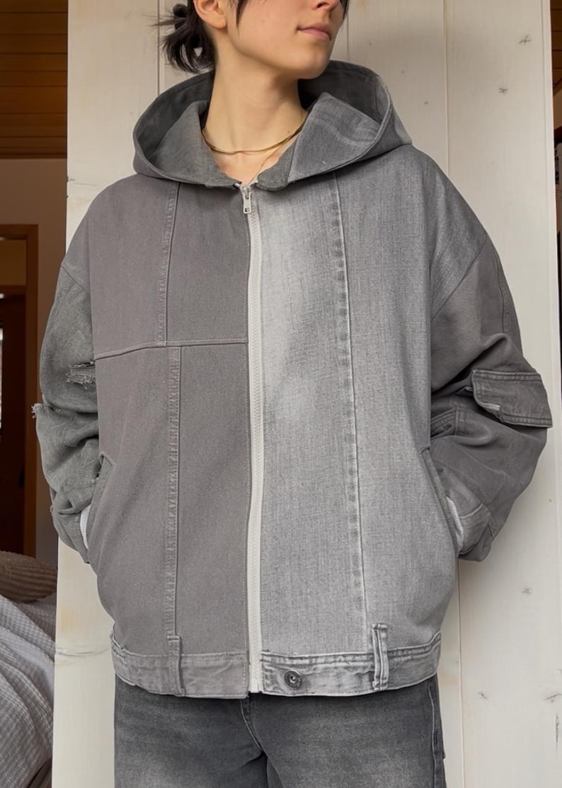 May include: A gray hooded jacket with a zip-up front. The jacket is a patchwork design, with different shades of gray denim. It has a hood, pockets, and a button closure at the waist. The jacket is worn with gray denim pants.