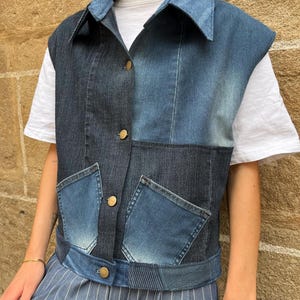 May include: A denim vest with a patchwork design in various shades of blue. The vest features a collar, two front pockets, and gold-colored buttons. The garment is worn over a white t-shirt and blue striped pants.