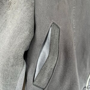 May include: Close-up of a gray denim jacket with a distressed, frayed section on the left shoulder. The jacket features a pocket with a gray and white striped lining. The stitching is visible, and the overall aesthetic is casual.