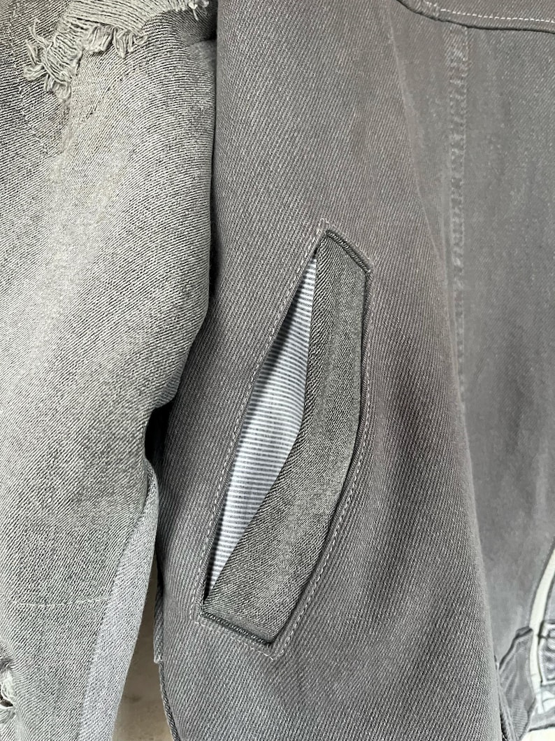 May include: Close-up of a gray denim jacket with a distressed, frayed section on the left shoulder. The jacket features a pocket with a gray and white striped lining. The stitching is visible, and the overall aesthetic is casual.