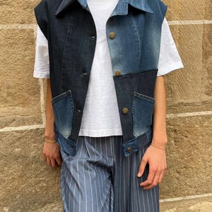 May include: A sleeveless denim vest in a patchwork design, featuring shades of blue. The vest has a collared neckline, button closures, and two front pockets. The person is wearing a white t-shirt and blue striped pants.