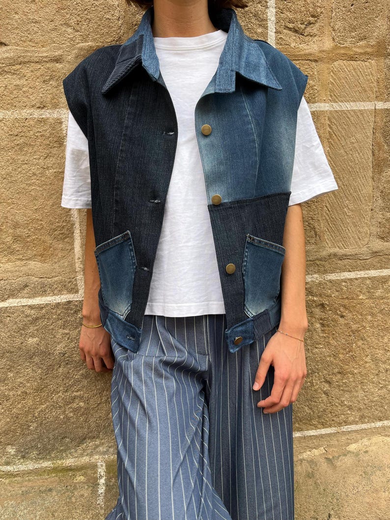 May include: A sleeveless denim vest in a patchwork design, featuring shades of blue. The vest has a collared neckline, button closures, and two front pockets. The person is wearing a white t-shirt and blue striped pants.