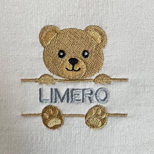 May include: White fabric with an embroidered teddy bear design. The bear is light brown with black eyes and a black nose. The word "LIMERO" is embroidered in gray below the bear.