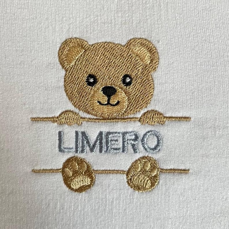 May include: White fabric with an embroidered teddy bear design. The bear is light brown with black eyes and a black nose. The word "LIMERO" is embroidered in gray below the bear.