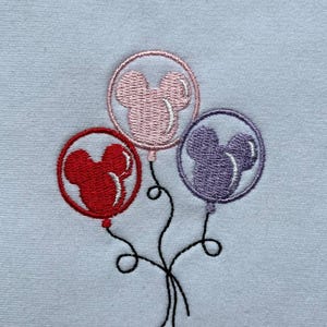 May include: Embroidered design featuring three balloons in red, pink, and purple, each shaped like a mouse head. The balloons are connected by black strings, creating a whimsical image. The background is a light blue fabric.