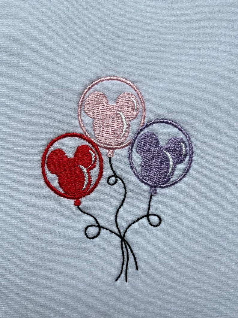 May include: Embroidered design featuring three balloons in red, pink, and purple, each shaped like a mouse head. The balloons are connected by black strings, creating a whimsical image. The background is a light blue fabric.