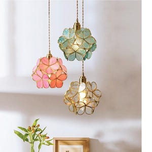 Puede incluir: Tres lámparas colgantes con pantallas en forma de flor en vidrio rosa, verde y transparente. Cada lámpara tiene un marco de metal dorado y un cable decorativo. Las lámparas cuelgan frente a una pared blanca.