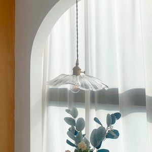 May include: A clear glass pendant light with a ruffled shade hangs from a brown cord. The light fixture has a brass-colored base and a visible lightbulb. The background includes a white curtain and a floral arrangement.