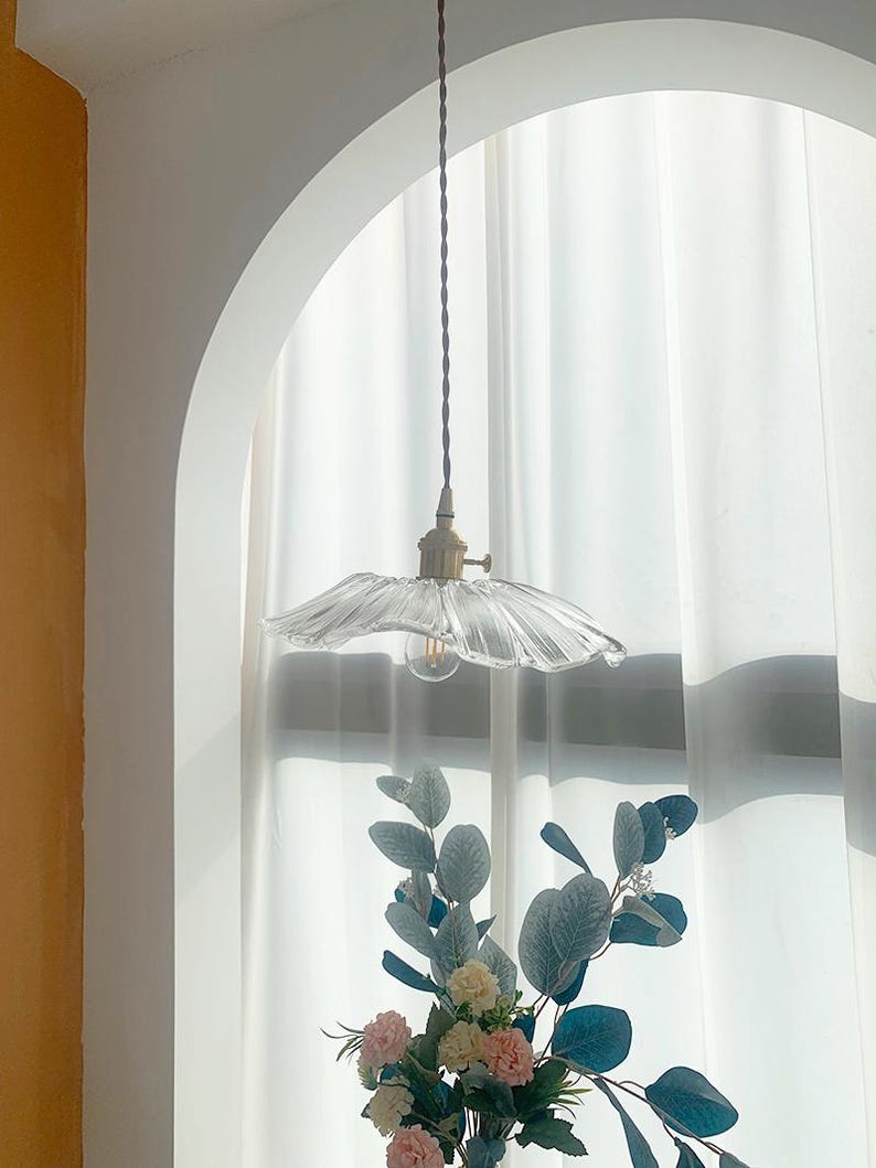 May include: A clear glass pendant light with a ruffled shade hangs from a brown cord. The light fixture has a brass-colored base and a visible lightbulb. The background includes a white curtain and a floral arrangement.