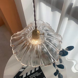 May include: Clear glass pendant light with a flower-shaped shade and a brass fixture. The light hangs from a brown twisted cord. A film clapperboard is on a white table below the light.