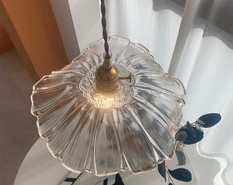 Op de afbeelding: Transparante glazen hanglamp met een bloemvormige kap en een messing armatuur. Het licht hangt aan een bruin gedraaid snoer. Een filmklapper ligt op een witte tafel onder het licht.