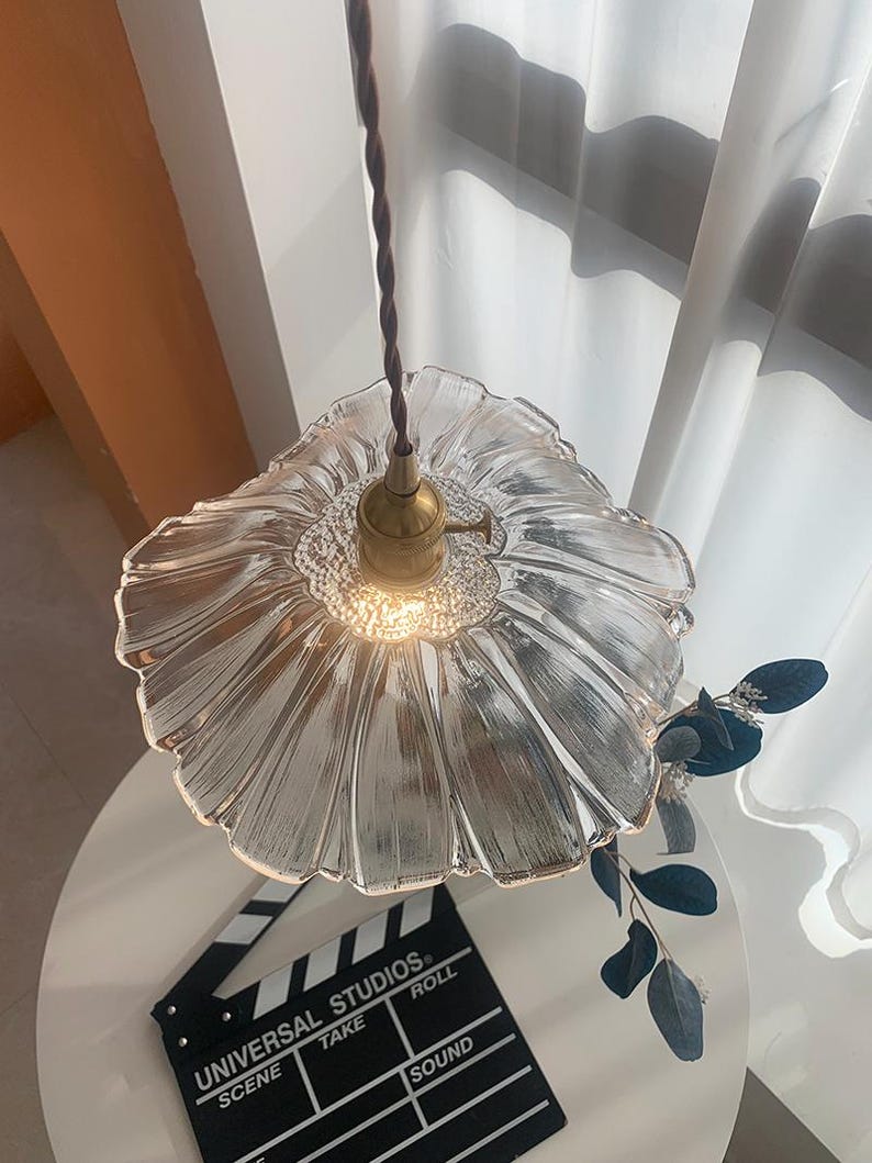 May include: Clear glass pendant light with a flower-shaped shade and a brass fixture. The light hangs from a brown twisted cord. A film clapperboard is on a white table below the light.