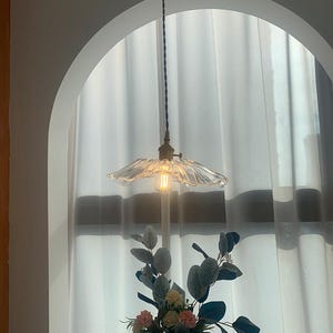 May include: A clear glass pendant light with a ruffled shade hangs from a brown cord. The light fixture is illuminated, casting a warm glow. Below, a vase holds a floral arrangement with pink, yellow, and green accents, set against a white curtain backdrop.