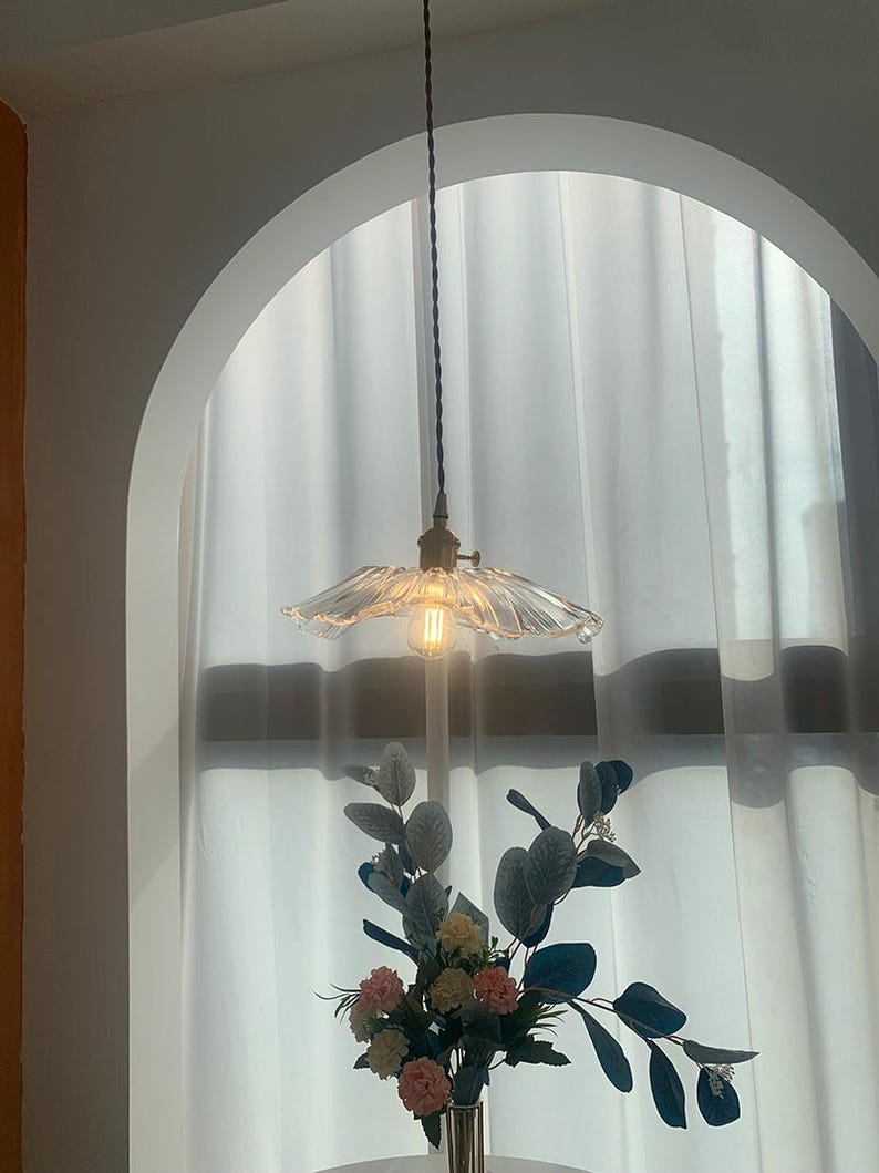 May include: A clear glass pendant light with a ruffled shade hangs from a brown cord. The light fixture is illuminated, casting a warm glow. Below, a vase holds a floral arrangement with pink, yellow, and green accents, set against a white curtain backdrop.