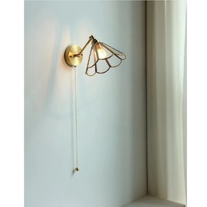 May include: A brass wall lamp with a pull chain. The lamp has a decorative, petal-shaped shade with a clear glass design. The lamp is mounted on a white wall.