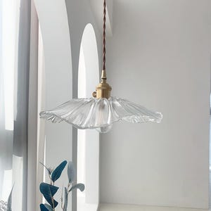 May include: A clear glass pendant light with a ruffled, flower-like shade. The light fixture has a brass-colored base and a brown twisted cord. The background is a white wall with an arched window.