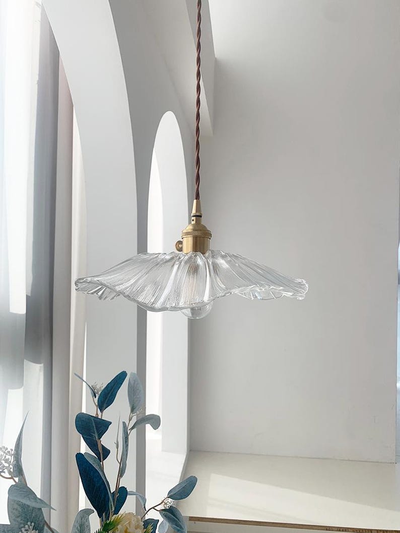 May include: A clear glass pendant light with a ruffled, flower-like shade. The light fixture has a brass-colored base and a brown twisted cord. The background is a white wall with an arched window.