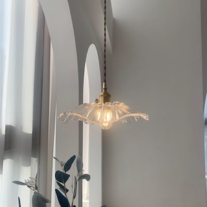 May include: A clear glass pendant light with a ruffled edge shade hangs from a brown cord. The light fixture has a brass-colored base and a visible lightbulb. The background features white walls and a glimpse of greenery.