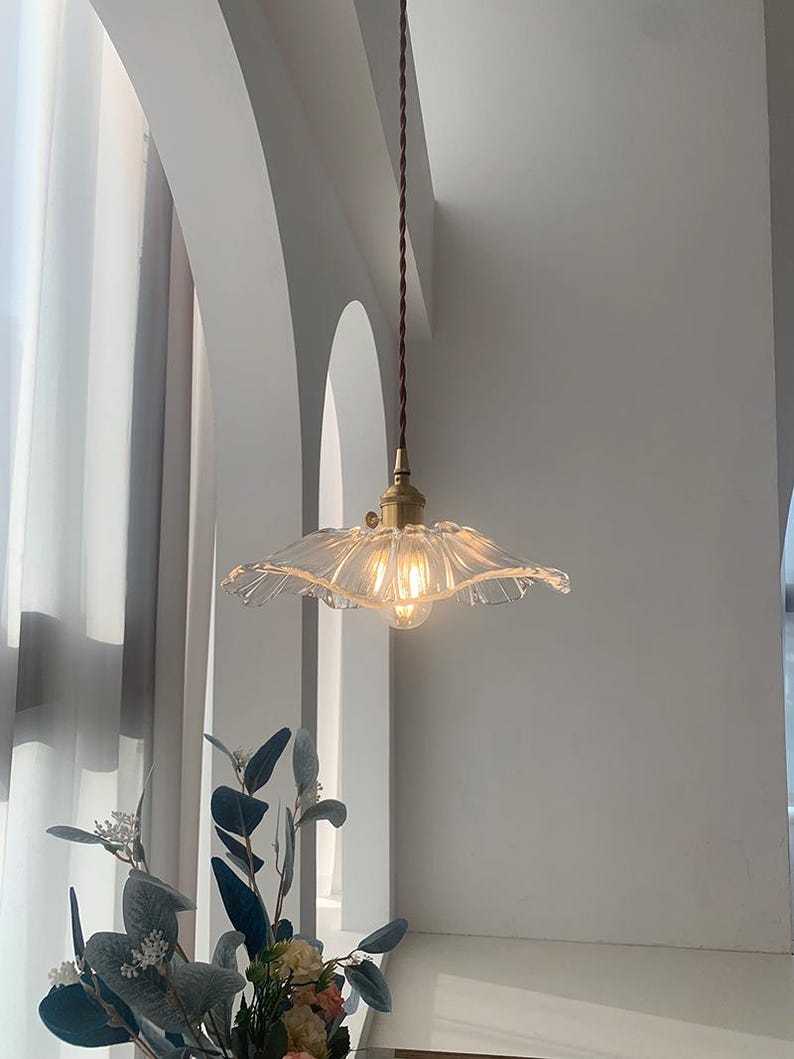 May include: A clear glass pendant light with a ruffled edge shade hangs from a brown cord. The light fixture has a brass-colored base and a visible lightbulb. The background features white walls and a glimpse of greenery.