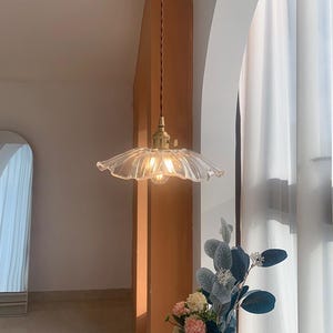 May include: A clear glass pendant light with a ruffled edge, hanging from a brown cord. The light fixture has a brass-colored base and is illuminated. The background includes a vase of flowers and a window with white curtains.